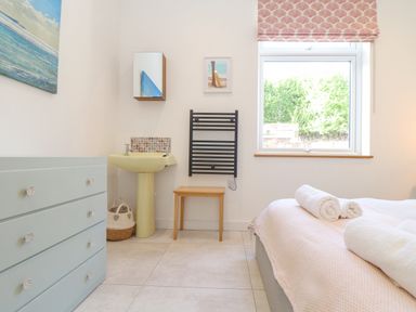 A bedroom with a bed with rolled towels a window a wooden stool a black wall rack a wall mirror a yellow sink a basket and a blue chest of drawers at Higher Bolenna in Perranporth