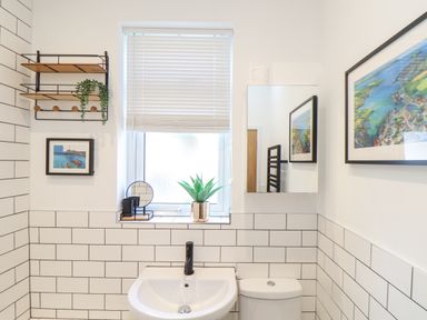 A bathroom with a sink, toilet, window with blinds, framed artwork, and a wall shelf with plants and accessories at Higher Bolenna in Perranporth