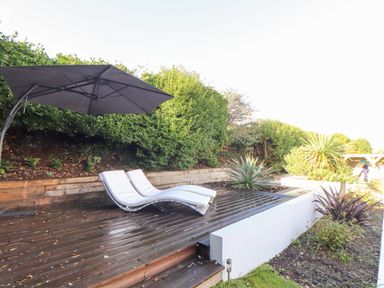 An outdoor deck with two white lounge chairs and a large black umbrella surrounded by plants and bushes at Higher Bolenna in Perranporth