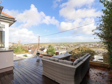 An outdoor wooden deck with wicker sofa and table overlooking a residential area and fields at Higher Bolenna in Perranporth