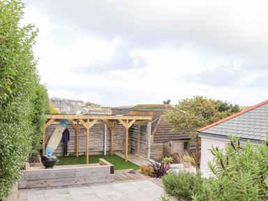 A backyard with a wooden pergola holding a surfboard and wetsuit next to bushes and garden beds at Higher Bolenna in Perranporth