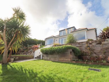 A garden with grass and tropical plants in front of a house with large windows at Higher Bolenna in Perranporth