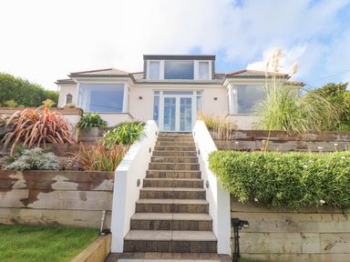 Exterior view of a house with a tiled staircase leading up to a white entrance door and landscaping on both sides at Higher Bolenna in Perranporth