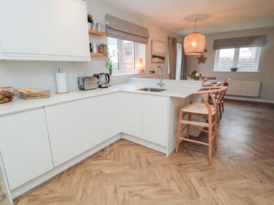 A kitchen with white cabinets, countertop, sink, wooden floor, bar stools, and dining area at Driftwood Sands in Beadnell