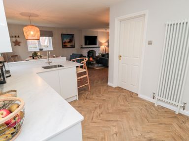 A kitchen with white countertops and wooden chair next to an open living area with blue sofas and a fireplace at Driftwood Sands in Beadnell