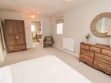 A bedroom with a wooden wardrobe a wooden dresser with a round mirror a chair and an open door showing a bathroom at Driftwood Sands Beadnell