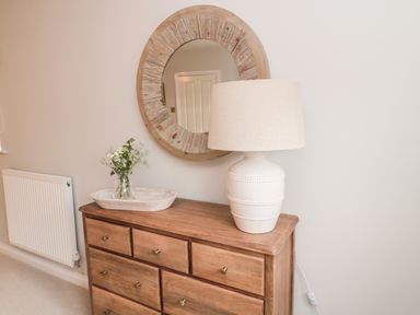A wooden dresser with a white table lamp and a small vase of flowers under a round wooden mirror at Driftwood Sands in Beadnell