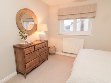 A bedroom with a wooden dresser a round wall mirror a table lamp a window and a bed at Driftwood Sands in Beadnell