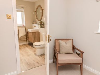 A wooden chair with a cushion next to a door leading to a bathroom with a toilet sink and round mirror at Driftwood Sands in Beadnell