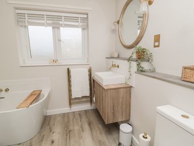 A bathroom with a white bathtub a wooden vanity with a sink a round mirror plants and a towel rack at Driftwood Sands in Beadnell
