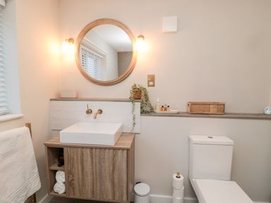 A bathroom with a wooden vanity and sink a round mirror above the sink a toilet and a small plant on a shelf at Driftwood Sands in Beadnell