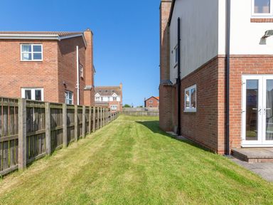 A grassy backyard area between brick houses with a wooden fence and clear sky at Driftwood Sands in Beadnell