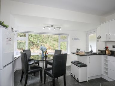 A kitchen with a round table and four black chairs next to a window and a white cabinet with a sink and trash bins at Ty Cerrig in Llanbedrog