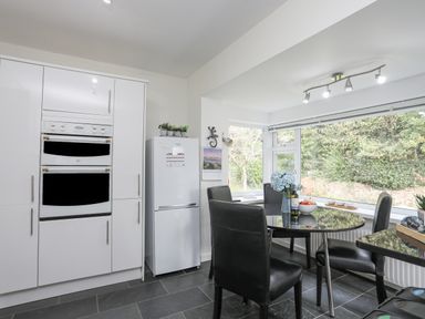 A kitchen with white cabinets a white refrigerator a round black dining table with black chairs and large windows at Ty Cerrig in Llanbedrog