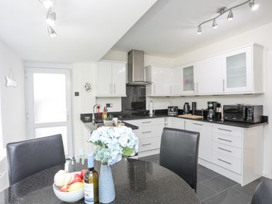 A kitchen with white cabinets black countertops a round table with flowers and fruit bowl at Ty Cerrig in Llanbedrog