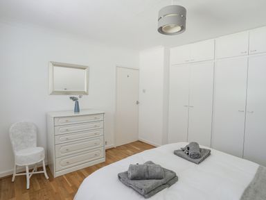 A bedroom with a bed, gray towels on the bed, a white chest of drawers with a vase, a wall mirror, a white chair, and built-in white wardrobes at Ty Cerrig in Llanbedrog