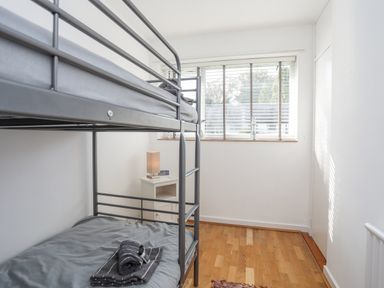 A bedroom with a metal bunk bed a small white bedside table with a lamp and clock and a window with blinds at Ty Cerrig in Llanbedrog