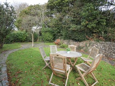 A garden with a round wooden table and six wooden chairs on grass near a stone wall in Ty Cerrig Llanbedrog