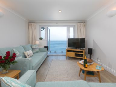 A living room with light blue sofas a wooden TV stand with a television a wooden coffee table with drinks and snacks and a view of the ocean through glass doors at Wavetop in Carbis Bay