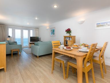 A living room and dining area with wooden table set for tea and sofas facing a TV with sea view at Wavetop in Carbis Bay