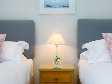 Two beds with gray headboards and white bedding with patterned pillows and a wooden nightstand with a lamp and flowers between them at Wavetop in Carbis Bay