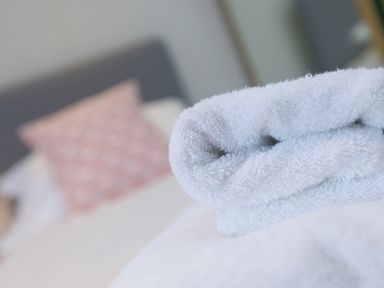 A folded white towel on a bed with a blurred pink patterned pillow and grey headboard in the background at Wavetop in Carbis Bay