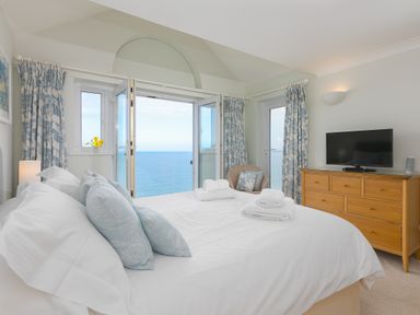 A bedroom with a large bed pillows and towels a wooden dresser with a TV a chair and windows with curtains overlooking the sea at Wavetop Carbis Bay