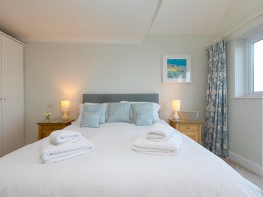 A bedroom with a double bed with blue patterned cushions two bedside tables with lamps folded white towels on the bed and a window with floral curtains at Wavetop in Carbis Bay