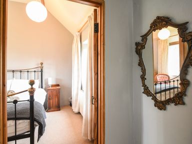 A bedroom with a metal bed frame bedside table and curtains seen through a doorway with an ornate wall mirror on the side at The Old Dairy in Whitchurch Canonicorum near Charmouth