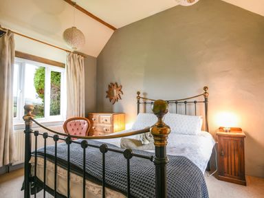 A bedroom with a metal bed frame a wooden dresser a wooden nightstand an upholstered chair and a window with curtains at The Old Dairy in Whitchurch Canonicorum near Charmouth