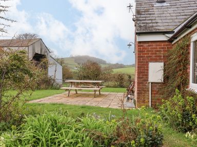 A garden with a picnic table on a stone patio next to a brick house and green hills in the background at The Old Dairy in Whitchurch Canonicorum near Charmouth