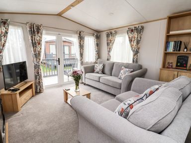 A living room with grey sofas a wooden TV stand a coffee table with flowers and a bookshelf at Silverdale View in East Heslerton near Sherburn