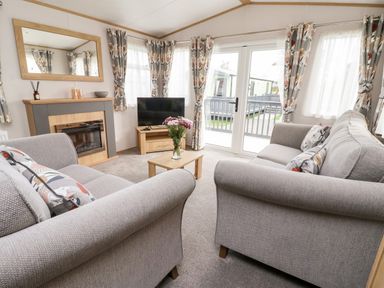 A living room with two gray sofas a wooden coffee table with flowers a fireplace a TV on a stand and large windows with patterned curtains at Silverdale View in East Heslerton near Sherburn