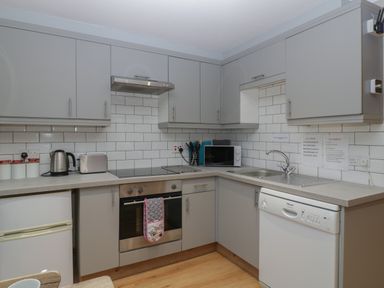 A kitchen with grey cabinets white tiled walls a stove dishwasher microwave kettle and toaster at Myrtle Cottage in St Brides-Super-Ely