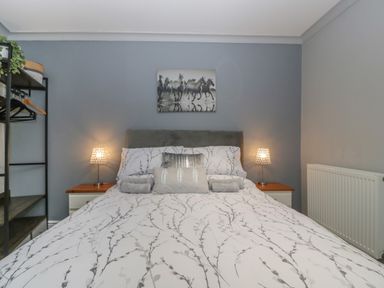 A bedroom with a bed in the center two bedside tables with lamps a wall radiator and an open shelving unit at Myrtle Cottage in St Brides-Super-Ely