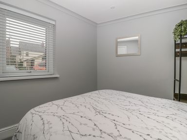 A bedroom with a bed covered in a white floral patterned duvet a window with blinds a mirror on the wall and a coat rack with plants at Myrtle Cottage in St Brides-Super-Ely