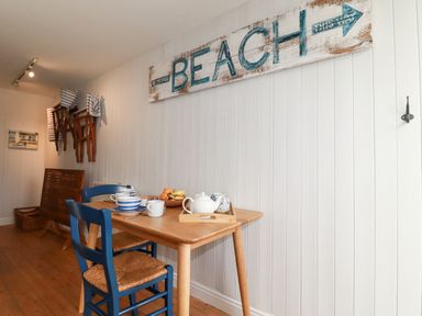 A dining area with a wooden table set with dishes and a tea set and blue chairs under a beach sign on a white wall at Waves End in Mevagissey