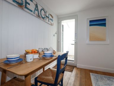 A dining area with a wooden table set with bowls and cups next to a door and a beach painting on the wall at Waves End in Mevagissey