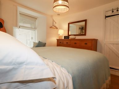 A bedroom with a bed covered in white and light blue bedding a wooden dresser with a mirror a lamp and a speaker in Waves End Mevagissey