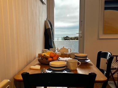 A dining table set with bowls cups fruit and a teapot near a door with a view of the sea at Waves End in Mevagissey