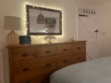 A wooden dresser with a lamp speaker diffuser and string lights around a mirror in a bedroom at Waves End in Mevagissey