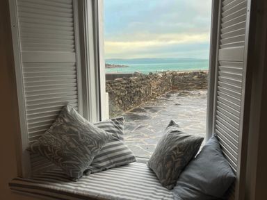 A window seat with striped cushion and four pillows facing a stone patio and ocean view at Waves End in Mevagissey