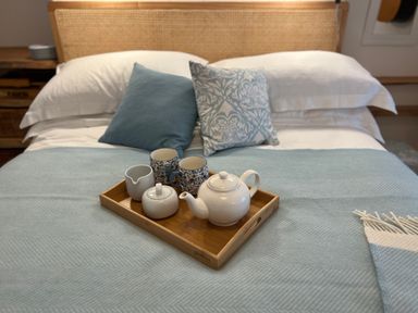 A bed with white pillows blue cushions and a wooden tray holding a teapot cups and a milk jug at Waves End in Mevagissey