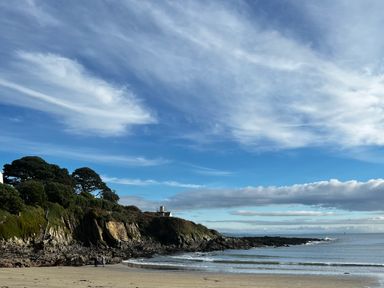 A rocky cliff with trees by a sandy beach and ocean under a partly cloudy sky at Waves End in Mevagissey