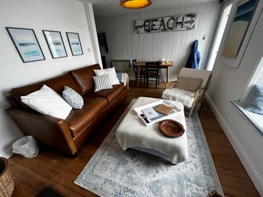 A living room with a brown leather sofa, armchair, ottoman, small dining table with chairs, and beach sign at Waves End in Mevagissey