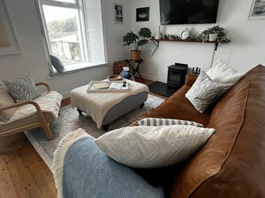 A living room with a brown sofa a wooden chair an ottoman with a tray a window and a wall-mounted television at Waves End Mevagissey