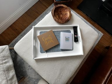 A white tray with a guest book a booklet and a remote control on a knitted ottoman in a living room at Waves End in Mevagissey