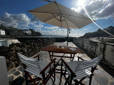 A patio with two striped chairs a wooden table with a tea set under a large umbrella overlooking a waterfront with buildings and hills at Waves End in Mevagissey