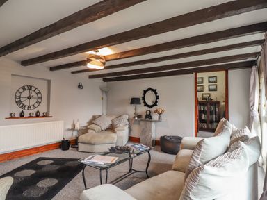 A living room with beams on the ceiling a large clock on the wall a sofa and armchair a glass coffee table and a doorway to another room at High Shaftoe in Ponteland