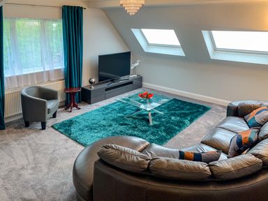 A living room with a curved leather sofa a glass coffee table a TV on a stand and teal curtains at The Gables Penthouse in Bournemouth